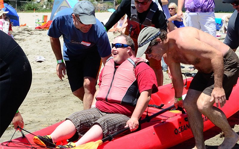 Volunteers carry a disabled person in a kayak for a ride in the ocean.