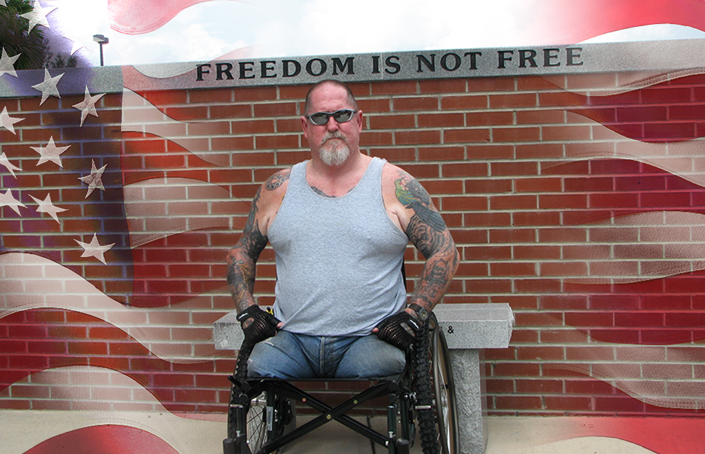 Image of a double amputee man in a wheelchair in front of a wall inscribed with "Freedom is not free." The American flag is laid over the image.