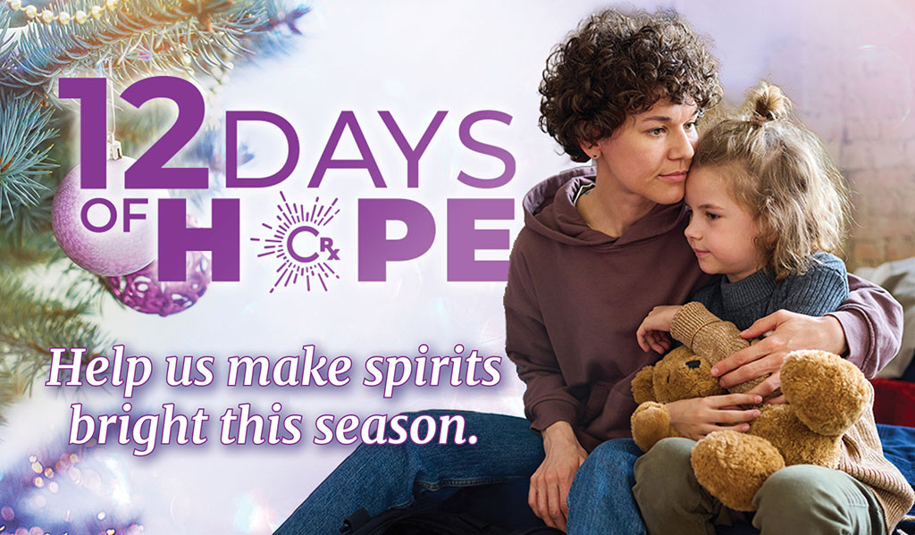 Image: A mother and child sit together on a cot with a sleeping bag on it. The child is holding a stuffed bear and the mother has her arm around the child. CharityRx 12 Days of Hope. 