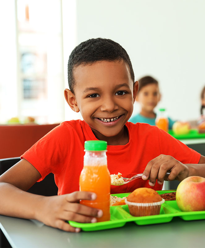 CharityRx cares about kids school lunch debt. A young boy smiling and sitting at a school lunch table with a tray of food in front of him.