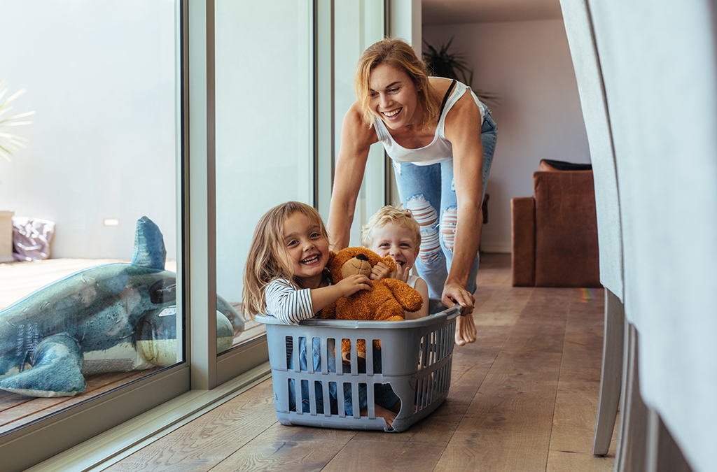 CharityRx Charity of the Month Domestic Violence. Image: A happy mother pushes her two small children along the floor in a laundry basket. They are laughing and enjoying the fun.