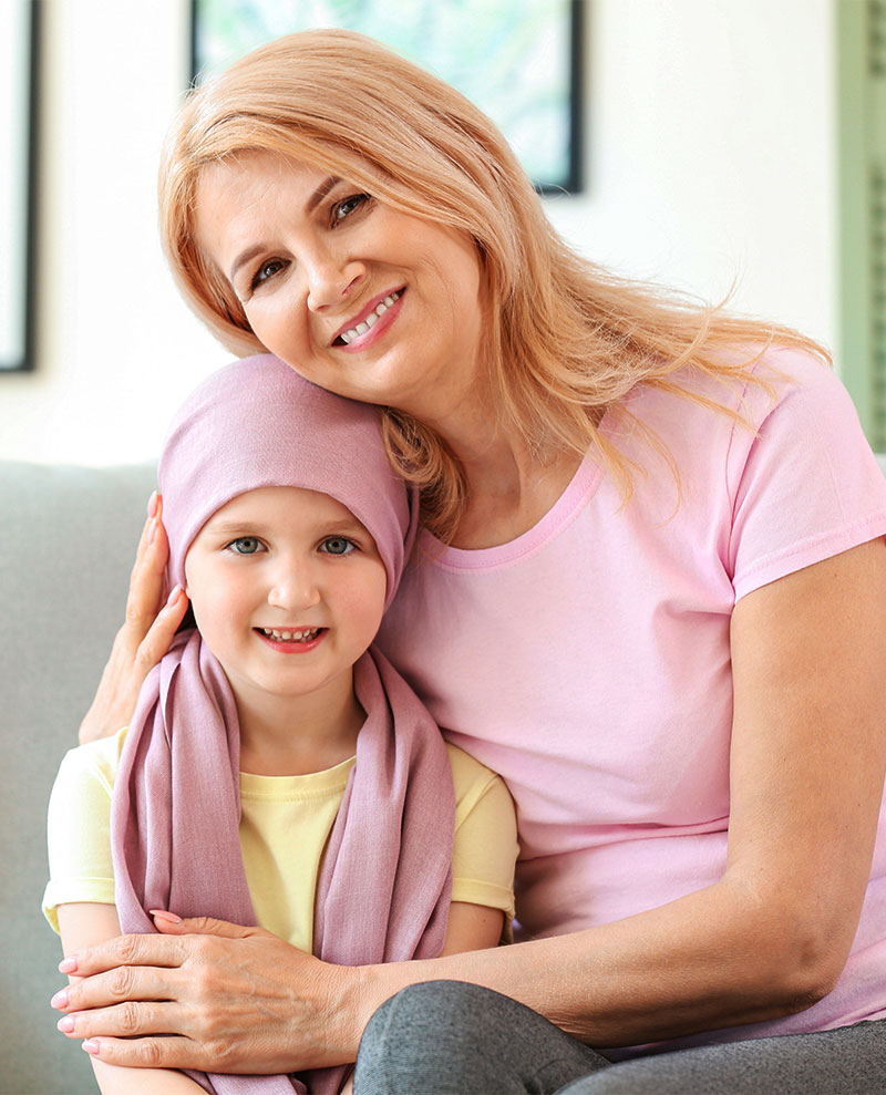 A young girl being embraced by a woman and sitting on a sofa. The young girl has a pink scarf over her head.