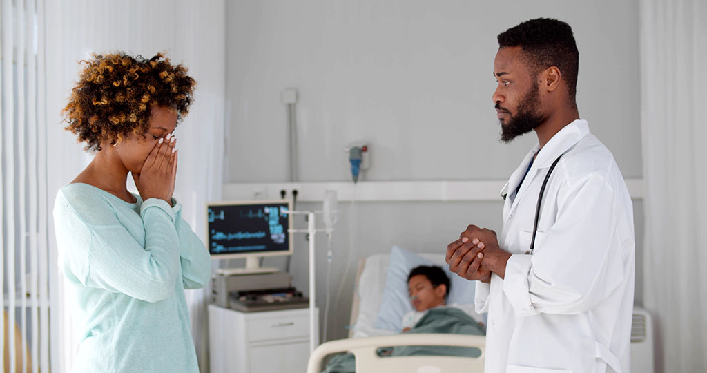 Image of a distraught Black mother and caring doctor talking in a hospital room. Behind them, a young Black boy is sleeping in a hospital bed, hooked up to monitors.