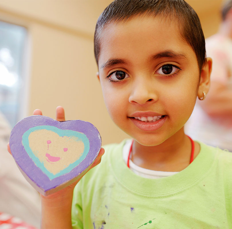 A young girl with shaved hair holding a hand-painted heart with a smiley face on it.