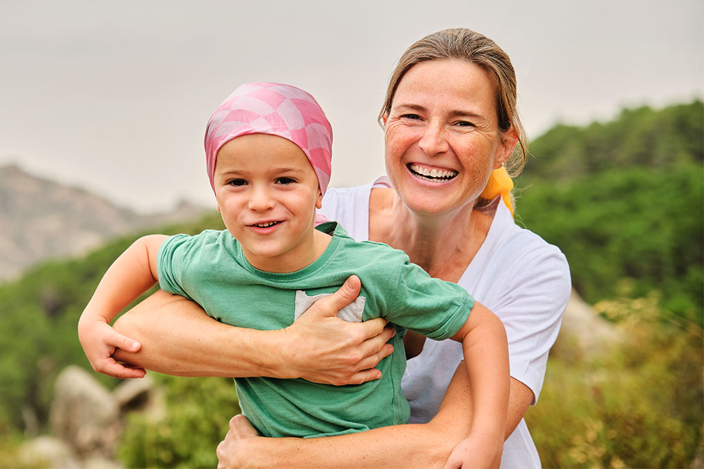 A smiling woman playing outside with a young child who is wearing a scarf tied tightly around their head.