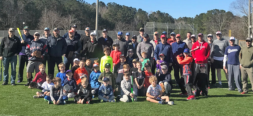 A large group of men and boys sitting on the grass at a park. Some are holding baseball bats and gloves.