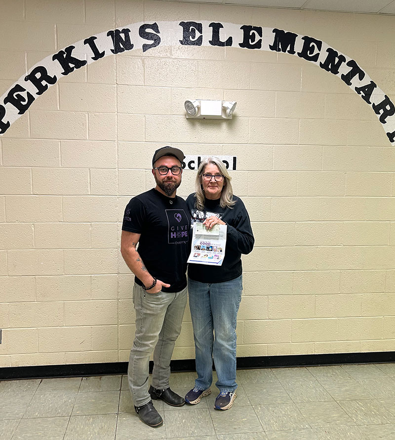 CharityRx cares about kids school lunch debt. A man and a woman stand side by side against a wall under letters that say Perkins Elementary. The woman is holding a check and a flyer.