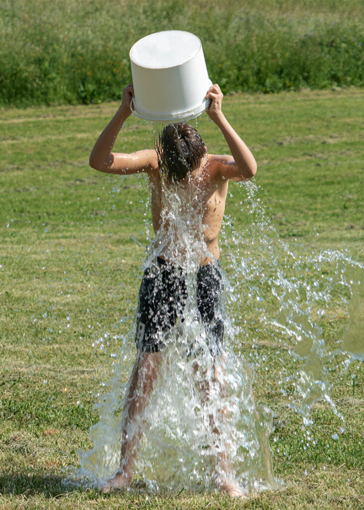Image of a young man standing in a grassy landscape, pouring a bucket of ice water over his head.