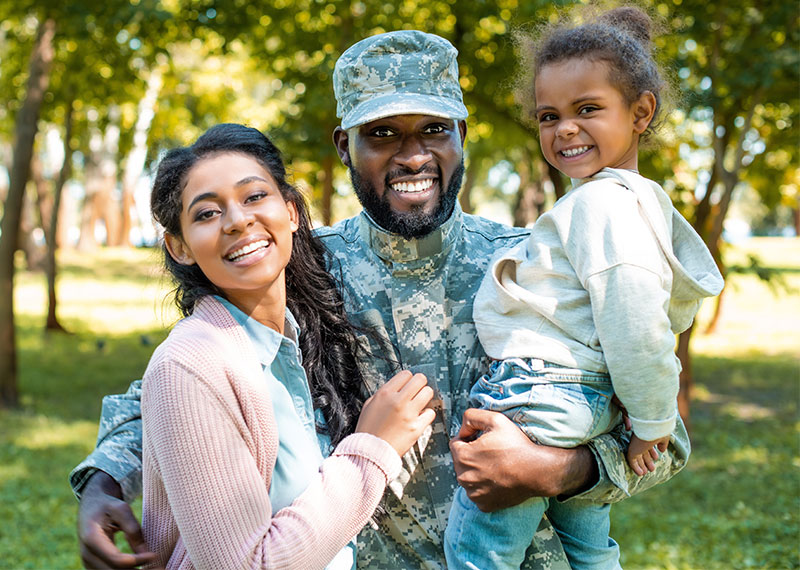 Image of a smiling, young, black family. The father, dressed in camouflage, standing in the center with his arm around his wife and young daughter in his arms.