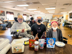 A middle aged man sits between two elderly residents behind a table at a care center. On the table are ingredients for an ice cream sundae party. CharityRx Reps Serve Nationwide on Founder’s Day