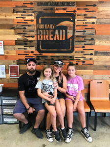 A family of four with two young girls sits smiling on a bench in front of a sign for Our Daily Bread Food and Resource Center. CharityRx Reps Serve Nationwide on Founder’s Day