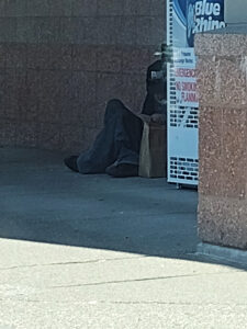 A homeless man sitting on the sidewalk, against a wall behind a propane tank return closet. A paper bag with snacks and water is next to him on the sidewalk. CharityRx Reps Serve Nationwide on Founder’s Day