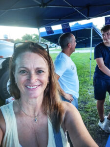 A smiling middle-aged woman stands in front of a man who is speaking with a young teenager under a pop-up shade tent. CharityRx Reps Serve Nationwide on Founder’s Day