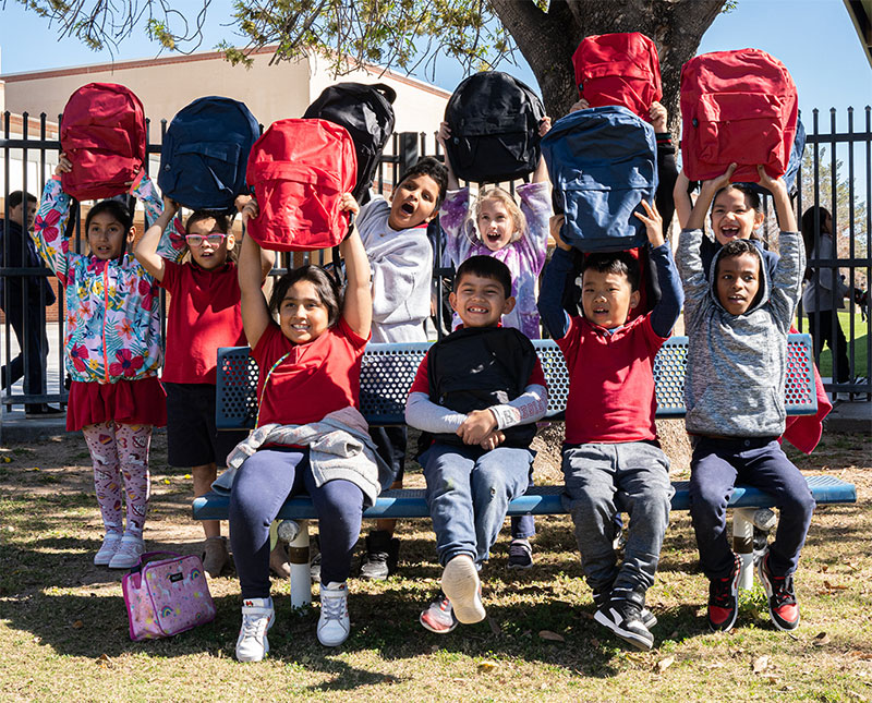 CharityRx Donates School Supplies. Image: A group of smiling young children outside a school. Each child holds up a backpack. 