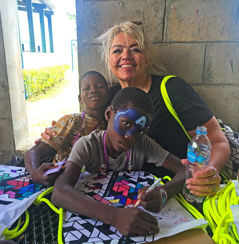 Meet CharityRx Rep, Carolyn Johnson. A smiling white woman with blonde hair sits with her arms around two young black boys at a table in front of a block wall. One boy has a Captain America mask painted on his face and is coloring a white backpack with markers.