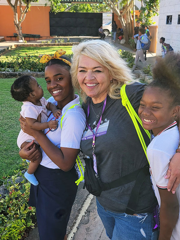 Meet CharityRx Rep, Carolyn Johnson. A smiling white woman with brown eyes and blonde hair stands with her arms around the shoulders of two smiling teenaged black girls. The older girl is holding a small black baby wearing a pink onesie.