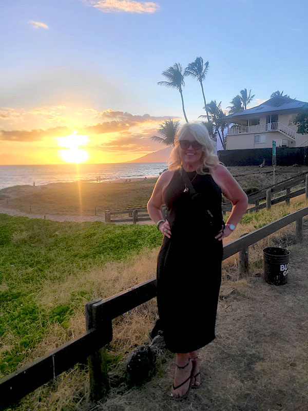 Meet CharityRx Rep, Carolyn Johnson. Image of a woman with blonde hair and sunglasses, wearing a sleeveless black dress. She is standing near a house and a fence in Hawaii with a view of the ocean at sunset.