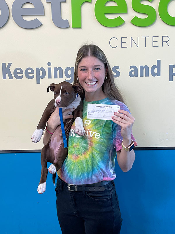 CharityRx Reps Rescue Shelter Pets with Donations. A young woman is wearing a tie-dyed t-shirt and holding a brown and white puppy in one arm while holding a check in the other hand.
