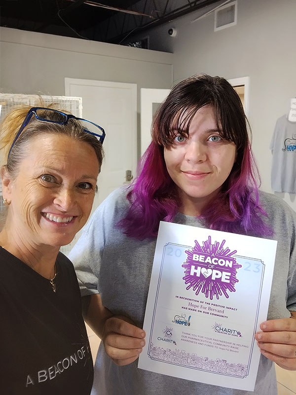 CharityRx Reps Rescue Shelter Pets with Donations. Two smiling women stand close together. One woman is blonde with a black t-shirt that reads "A Beacon of Hope." The second woman has medium-length dyed hair that goes from dark brown to pink to purple. This woman is holding a Beacon of Hope Award certificate.