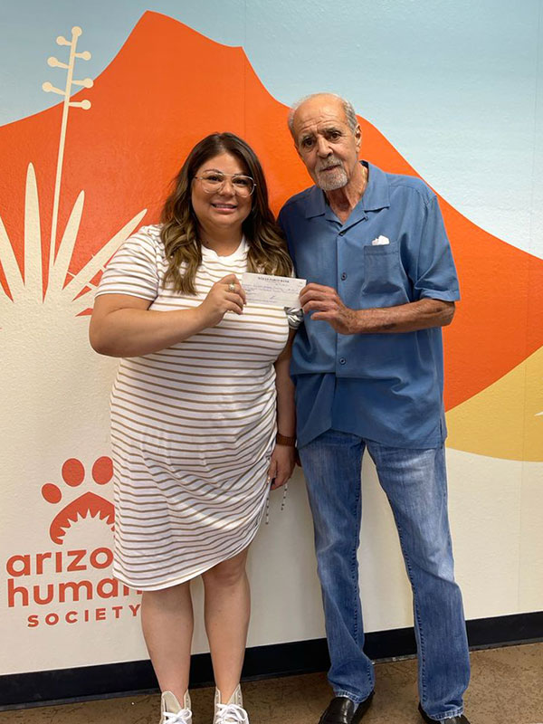 CharityRx Reps Rescue Shelter Pets with Donations. A woman and man stand together against a painted mural of red rocks and yucca plants in orange, yellow, and beige. They are holding a check between them and smiling. The Arizona Humane Society logo is behind them on the wall.