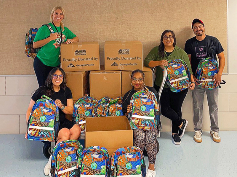 CharityRx Donates School Supplies. Image: A man and four women stand next to nine cardboard boxes filled with backpacks. Each person is smiling and holds a colorful backpack.