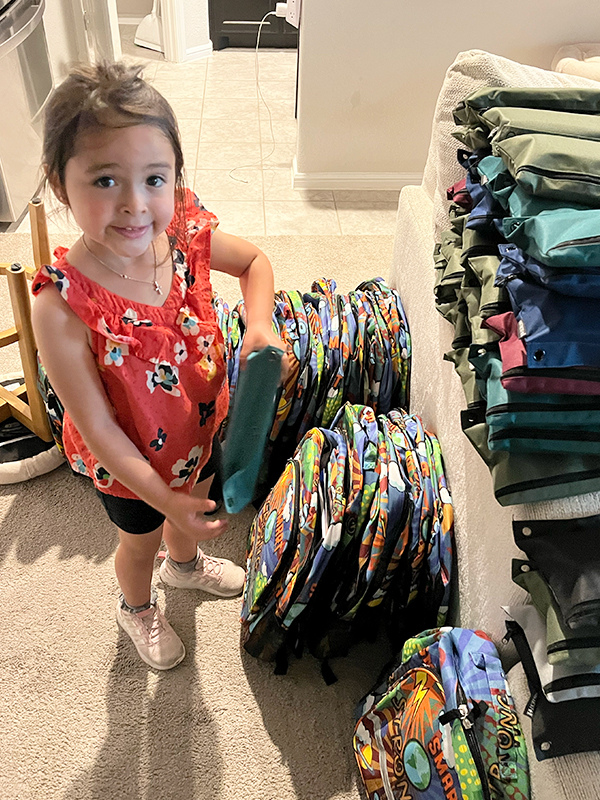 Image: A young girl in a red flowered tank top smiles at the camera and holds a pencil bag full of school supplies. Two stacks of empty backpacks are arranged at her feet.
