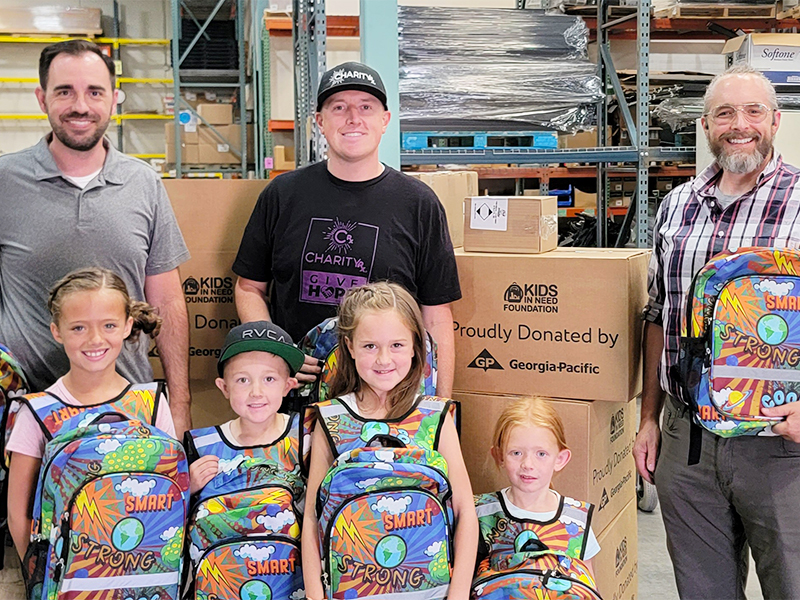 CharityRx Donates School Supplies. Image: Three men and four children stand smiling in front of several cardboard boxes that read "Kids In Need Foundation proudly donated by Georgia Pacific Inc." The children are wearing colorful backpacks on their chests.