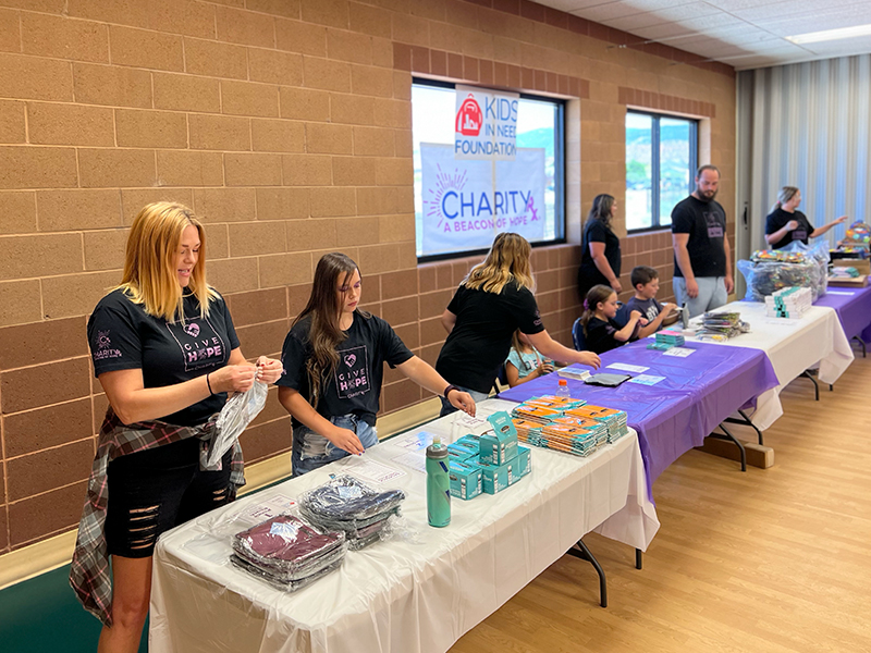 Image: A row of adults and children stand behind long tables, packing school supplies into backpacks.