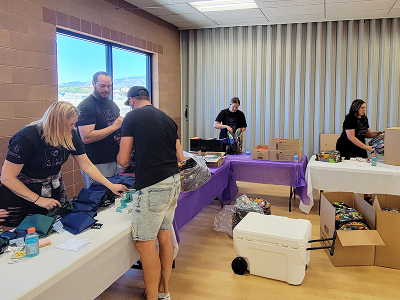 Image: Two men and three women pack school supplies into backpacks, check to make sure they are correct, and pack them into boxes.