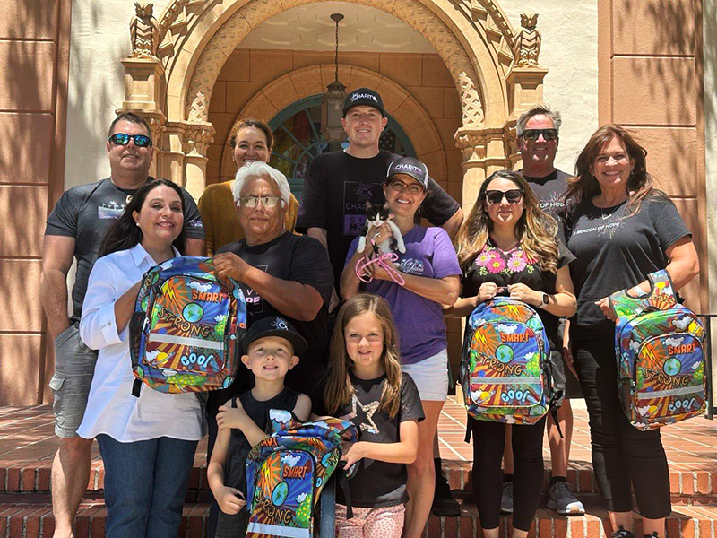 CharityRx Donates School Supplies. Image: A group of people stands together on the steps of a school, holding colorful backpacks.