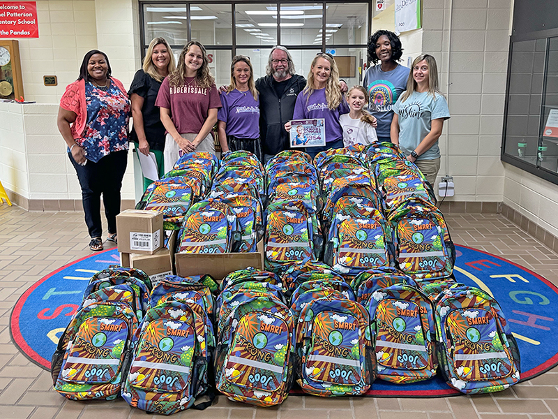 CharityRx Donates School Supplies. Image: A group of people stand behind 100 colorful backpacks arranged on the floor at their feet.