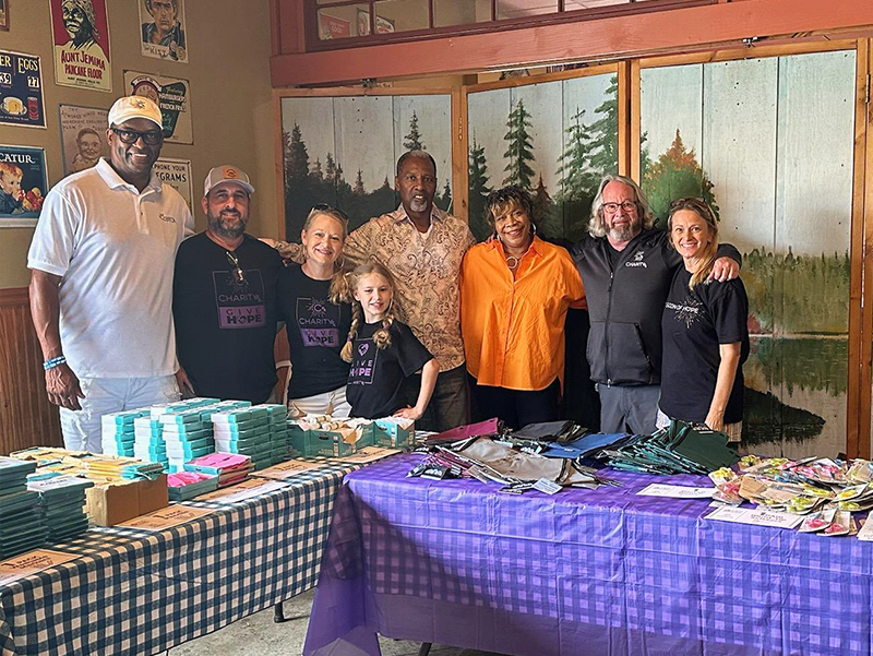 CharityRx Donates School Supplies. Image: A group of smiling people stand shoulder-to-shoulder behind tables with stacks of school supplies.