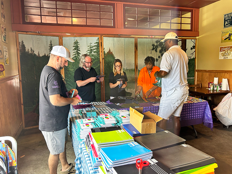 Image: Three men and two women stand in room with long tables covered in stacks of school supplies.