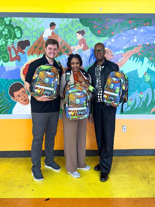 CharityRx Donates School Supplies. Image: Two men and a woman stand inside Chester Upland School of Arts school before a mural holding colorful backpacks. 