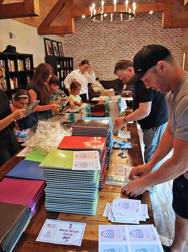 Image: A group of people stand on either side of a long wooden table, working to pack items into backpacks for children in need.