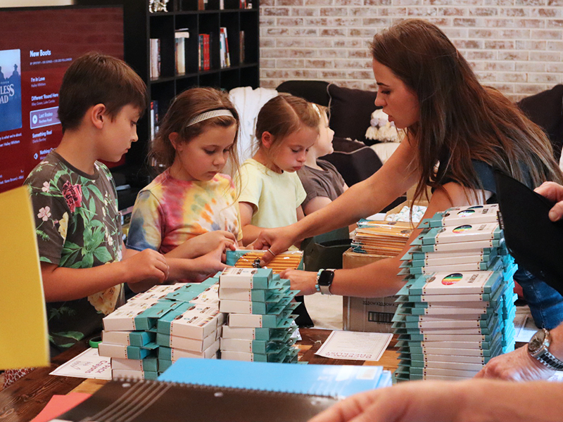 Image: Four children and one woman work together to stuff school supplies into backpacks at a table covered in supplies.