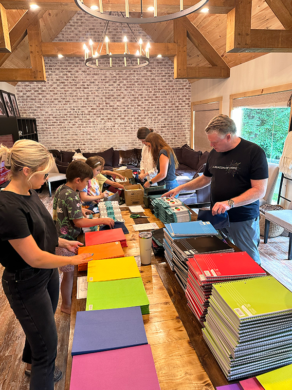 Image: A long wooden table is piled with stacks of colorful folders, notebooks, markers, and other school supplies. Several people stand on either side, organizing the piles.