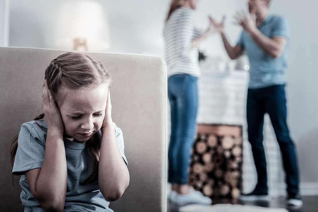 CharityRx Charity of the Month Domestic Violence. Image: a young white girl sits behind a sofa with her hands over her ears. In the background, her parents are fighting.