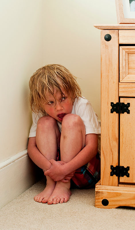 CharityRx Charity of the Month Domestic Violence. Image: A young boy with dirty hair and a fearful expression hides in the corner between a cabinet and the wall. He is peeking around the corner of the cabinet.