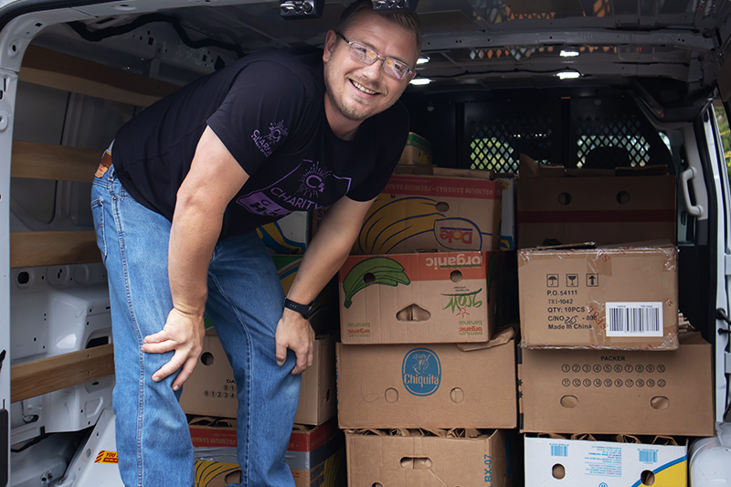 CharityRx Founder's Day 2023: The Power of Giving Back. Image: A smiling, middle-aged man wearing glasses, a black t-shirt and jeans looks out from the back of a van full of boxes.