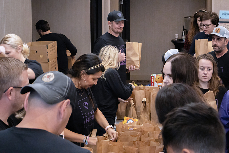 CharityRx Founder's Day 2023: The Power of Giving Back. Image: A group of people stand on either side of long tables stacked with brown paper bags and food items.