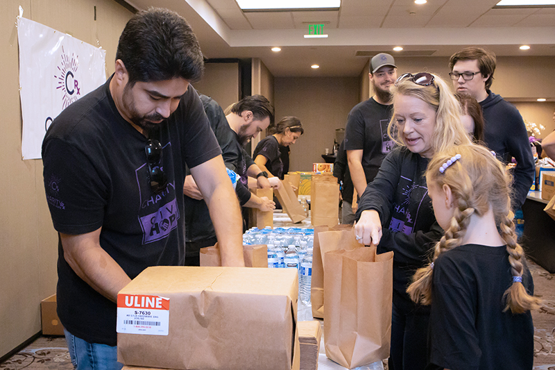 CharityRx Founder's Day 2023: The Power of Giving Back. Image: A group of people stand on either side of long tables stacked with brown paper bags and food items.