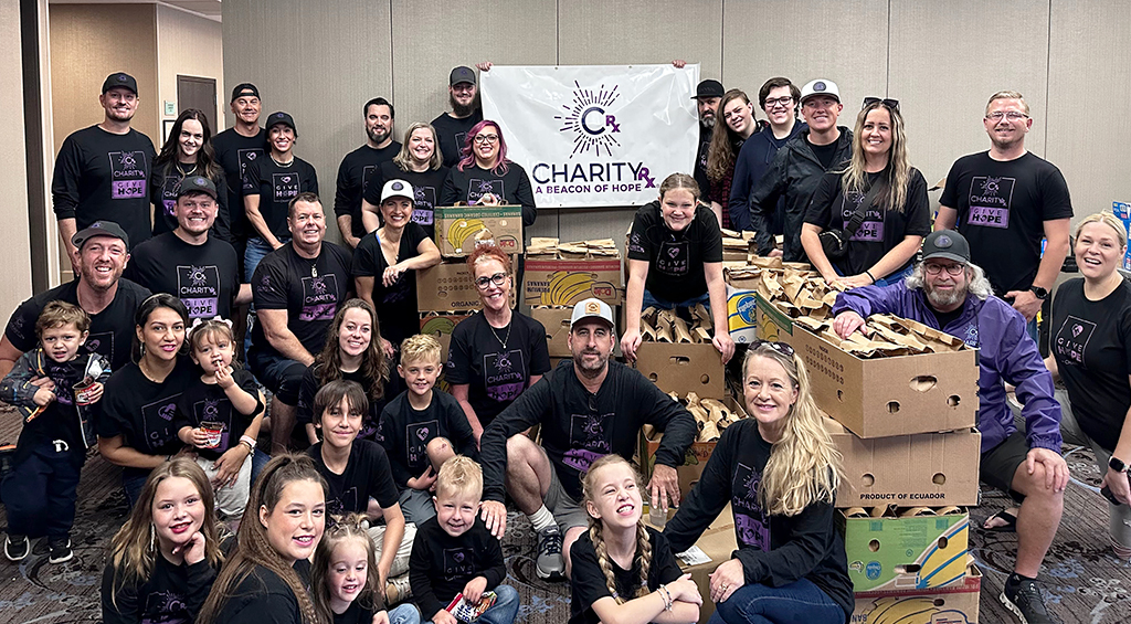 CharityRx Founder's Day 2023: The Power of Giving Back. Image: A large group of smiling people, adults and children, sit together around several stacks of boxes full of brown paper bags. All people are wearing black t-shirts with a purple "CharityRx Give Hope" logo.