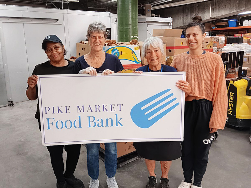 CharityRx Founder's Day 2023: The Power of Giving Back. Image: Four women of varying age and appearance stand inside a warehouse behind a large sign that reads "Pike Market Food Bank."
