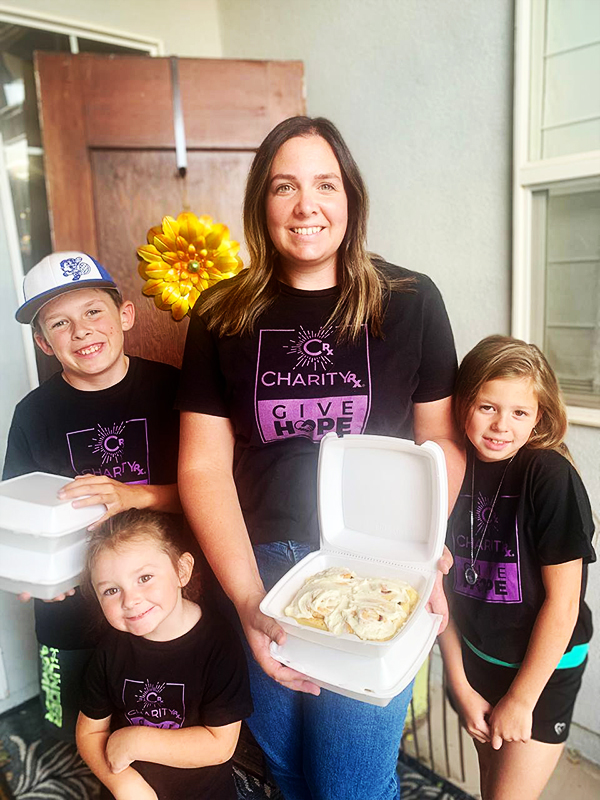 Image of a smiling woman holding a box of cinnamon rolls and standing in the midst of three young children who are also holding boxes. CharityRx Founder's Day of Service 2023