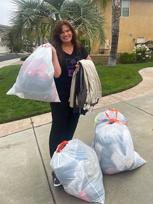 Image of a woman standing outside on a sidewalk, in front of a palm tree. She is holding a white trash bag full of clothes to be donated and has several pairs of pants slung over one arm. Three more bags of clothing sit on the ground at her feet. CharityRx Founder's Day of Service 2023