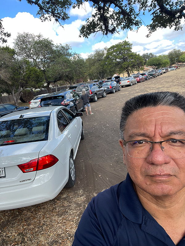 CharityRx Founder's Day 2023: The Power of Giving Back. Image: A middle-aged Latino man stands next to a long line of cars at a park.
