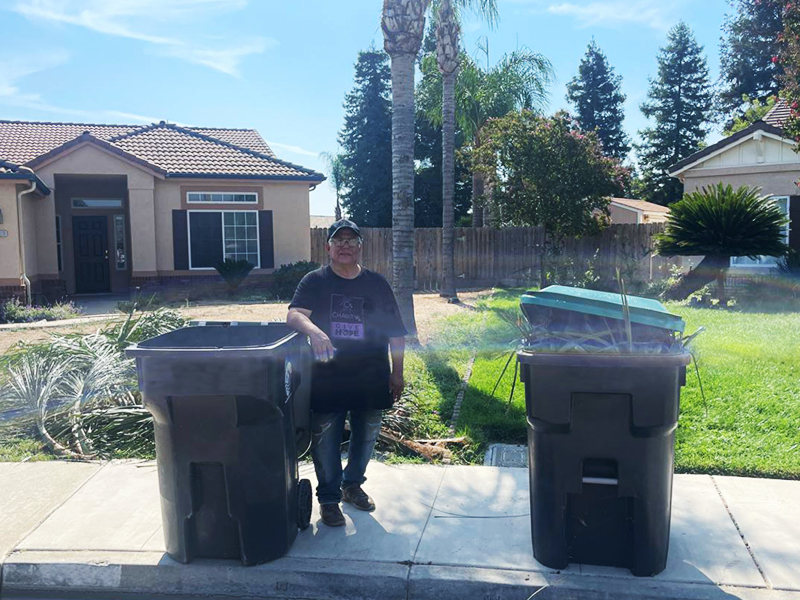 CharityRx Founder's Day 2023: The Power of Giving Back. Image: A middle-aged Latino man stands in front of two palm trees, between two garbage cans full of palm leaves.
