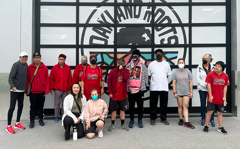 Image of a group of parents and their disabled children standing in front of a large window with the Oakland Roots logo on it. CharityRx Founder's Day of Service 2023