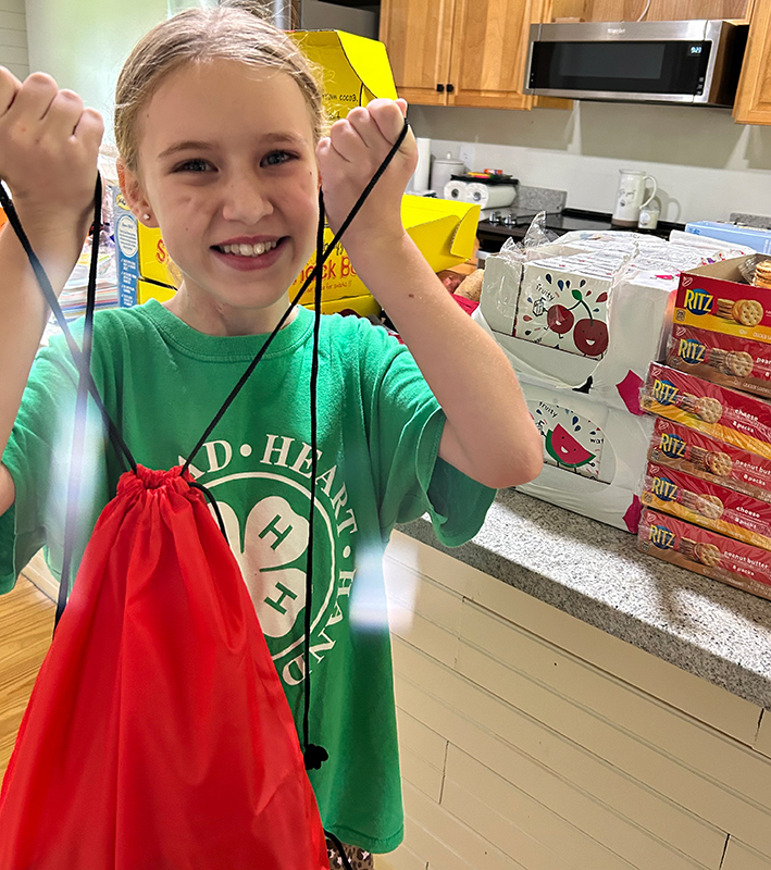 CharityRx Founder's Day 2023: The Power of Giving Back. Image: A smiling young, blond girl is standing in a kitchen, holding up a red string backpack. Several boxes of snacks are stacked on the counter behind her.
